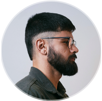 Foto de João Victor Beato de perfil lateral direito, usando óculos e camisa verde, barba e fundo branco acinzentado.